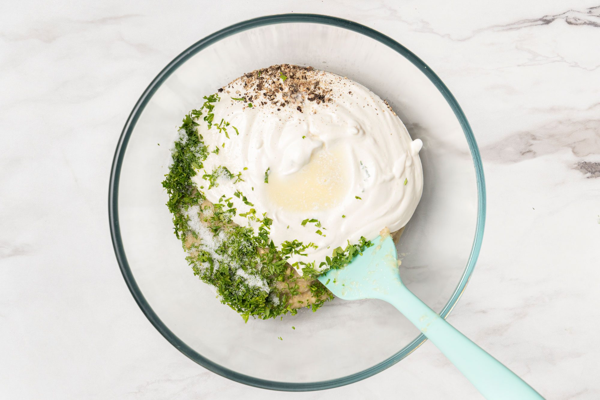 Overhead shot of a glass bowl filled with sour cream, chopped herbs, black pepper, salt, and lemon juice, with a light blue spatula resting inside on a marble surface;