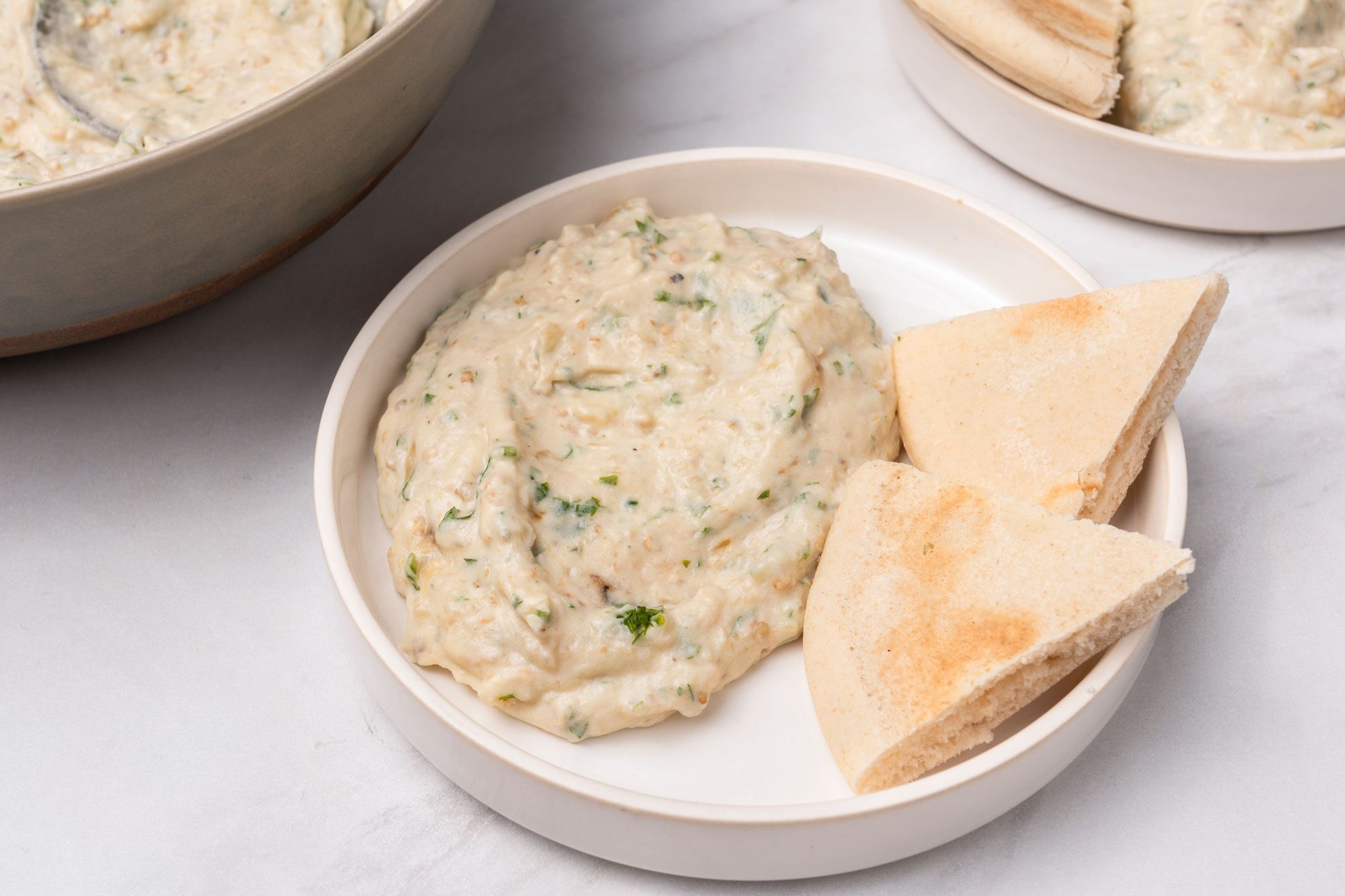 3/4 angle view shot of Mediterranean Eggplant Dip served with flat braeds on white plates; all set on a marble surface;