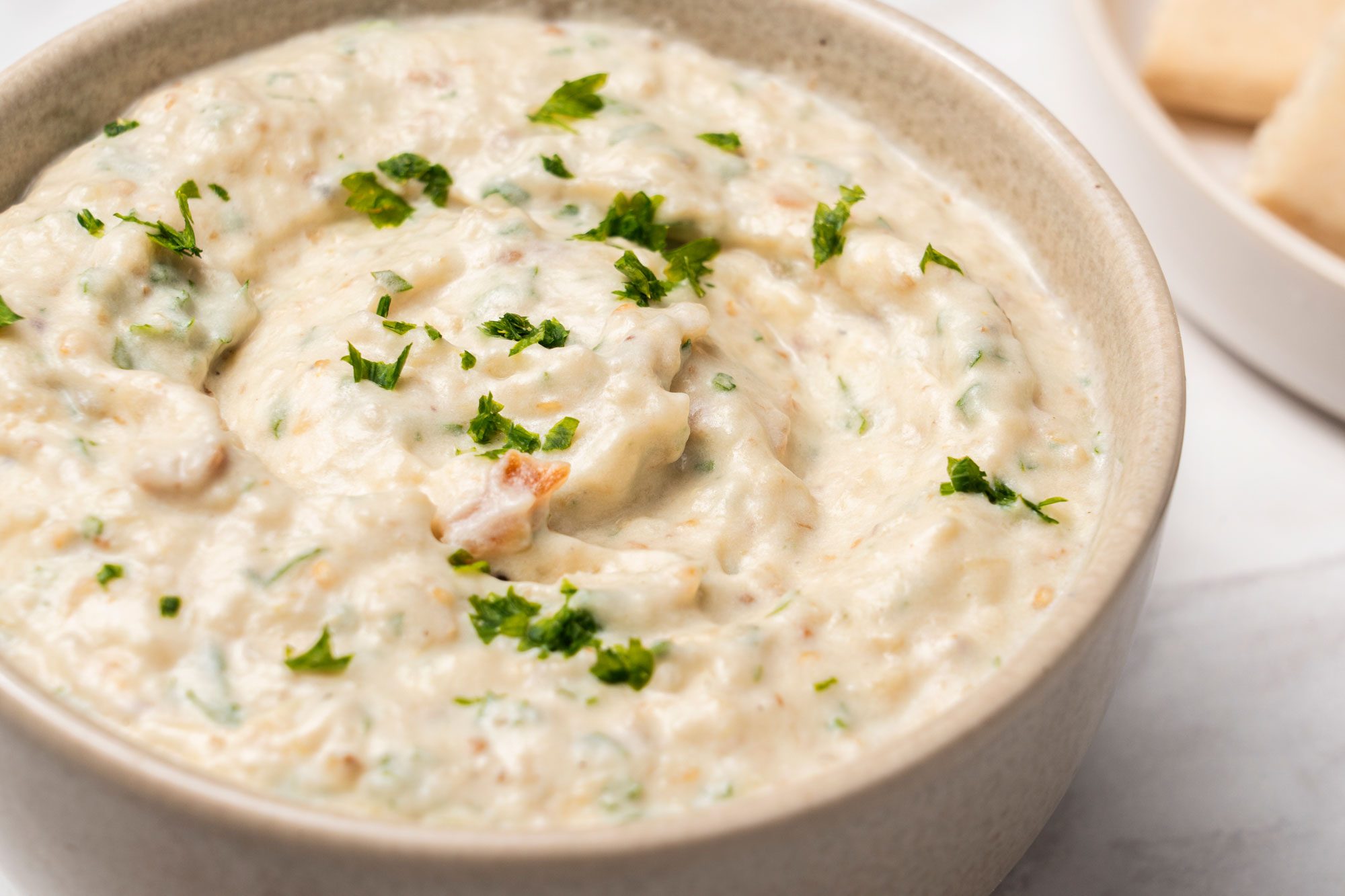 Close view shot of Mediterranean Eggplant Dip in a gray bowl; garnuish with chopped herb; served with flat breads on a wooden tray is visible in the background; all set on a marble surface;