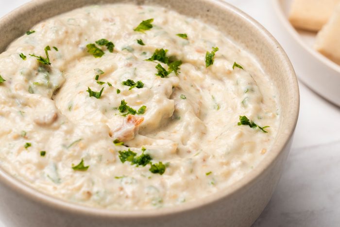 Close view shot of Mediterranean Eggplant Dip in a gray bowl; garnuish with chopped herb; served with flat breads on a wooden tray is visible in the background; all set on a marble surface;