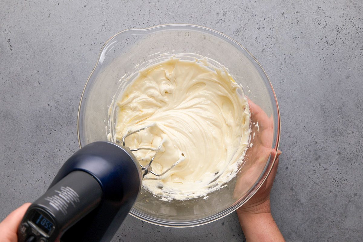 overhead shot of a hand holds a glass bowl filled with creamy batter while using a black electric hand mixer to blend the mixture on a gray countertop