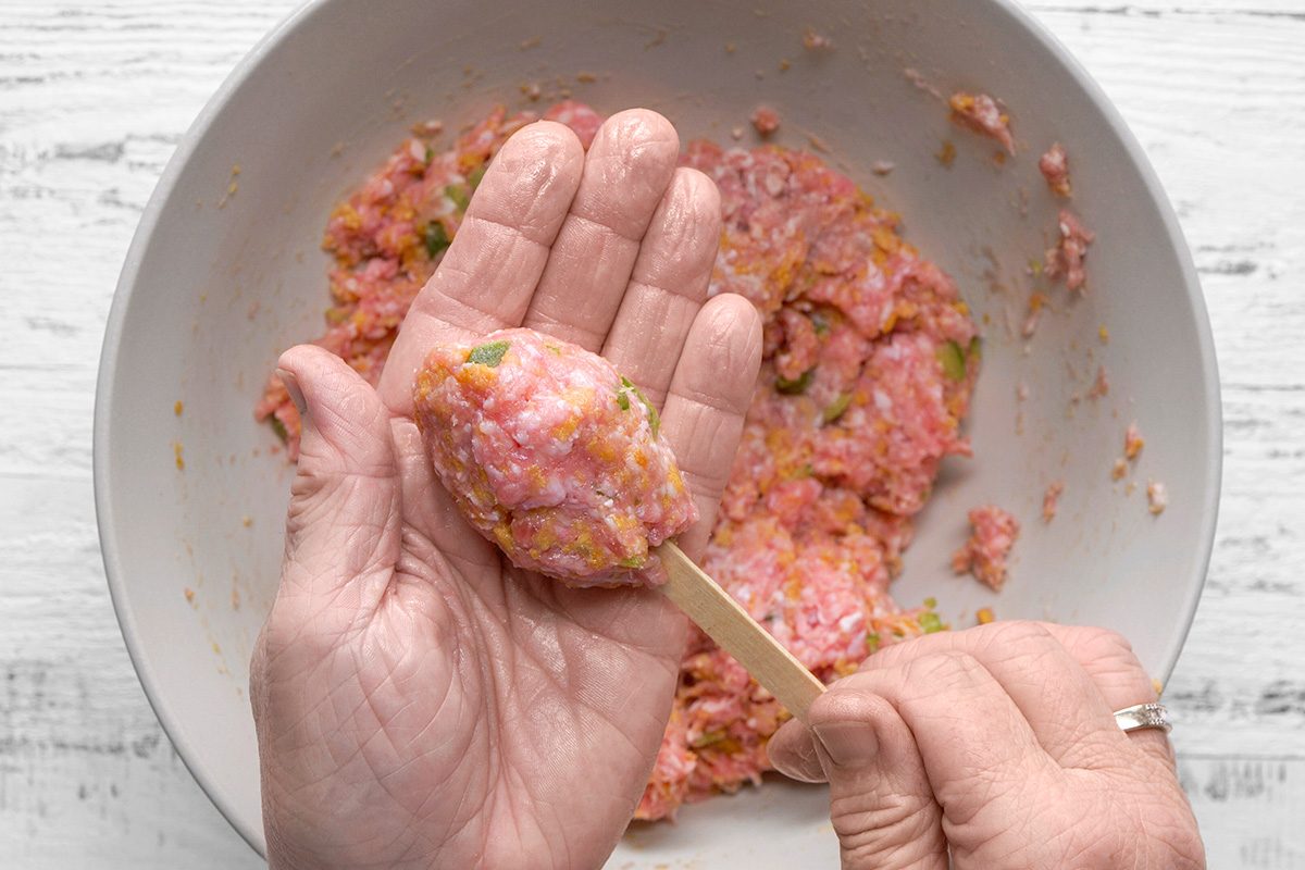 A person shaping ground meat mixture around a wooden stick over a bowl of the same mixture, preparing food on a light surface.