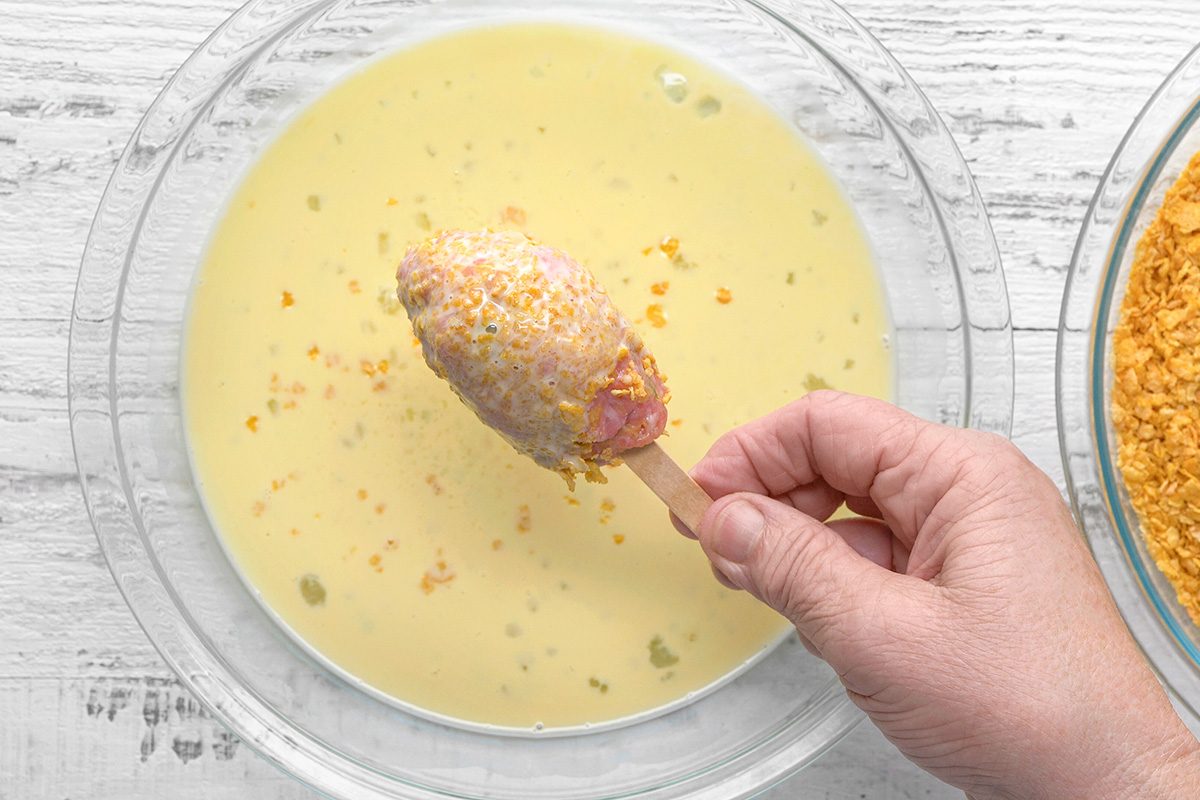 A hand dips a corn dog on a stick into a bowl of pale yellow batter. Another bowl with crushed coating is visible beside it, all set on a white wooden surface.