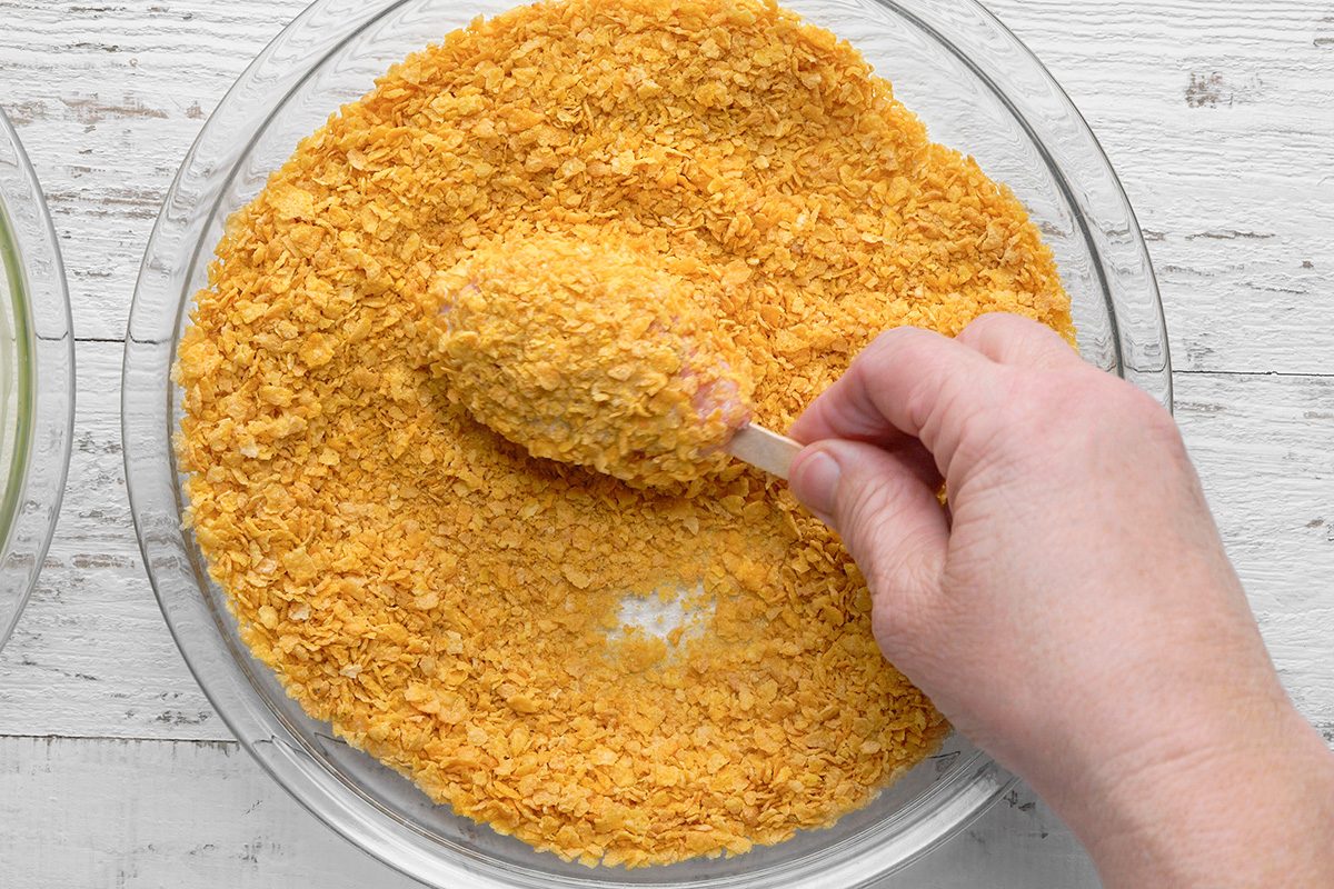 A hand dips a piece of food, likely chicken, into a bowl of crushed cornflakes or breadcrumbs, coating it evenly. The bowl sits on a white wooden surface.
