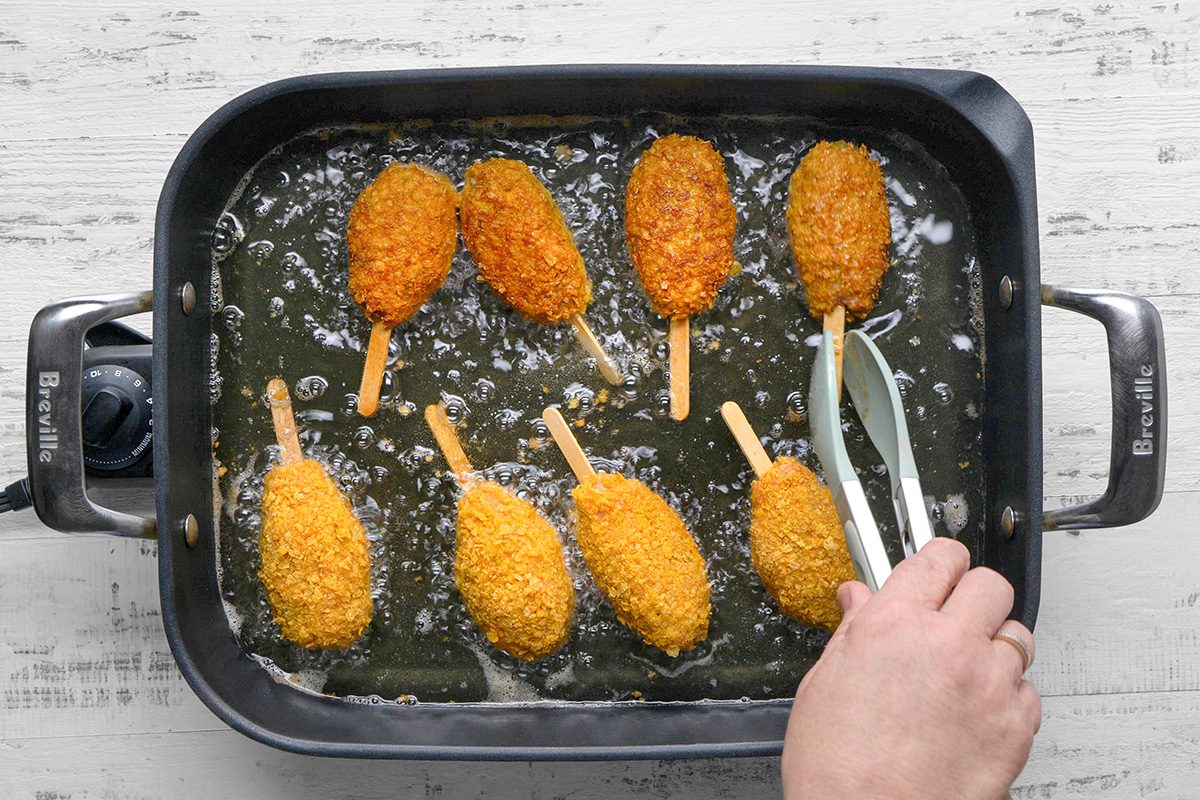 A hand uses tongs to flip breaded corn dogs frying in hot oil inside a large black electric skillet on a light-colored surface.