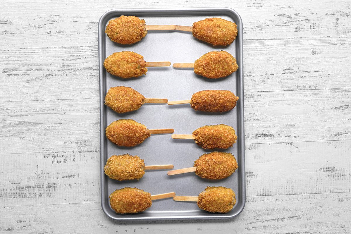 A baking tray with ten breaded, oval-shaped snacks on wooden sticks arranged in two columns on a white wooden surface.
