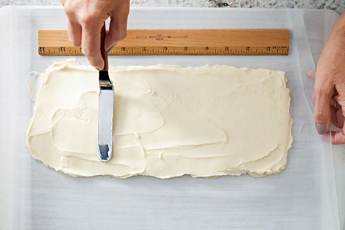 Overhead shot of a hand using an offset spatula to spread white icing evenly on a rectangular sheet of parchment paper; with a wooden ruler placed along the top edge for measurement;