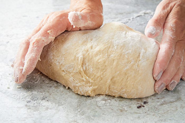 Overhead shot of two hands kneading a ball of dough on a floured countertop; with flour dusted across the hands and surrounding surface.