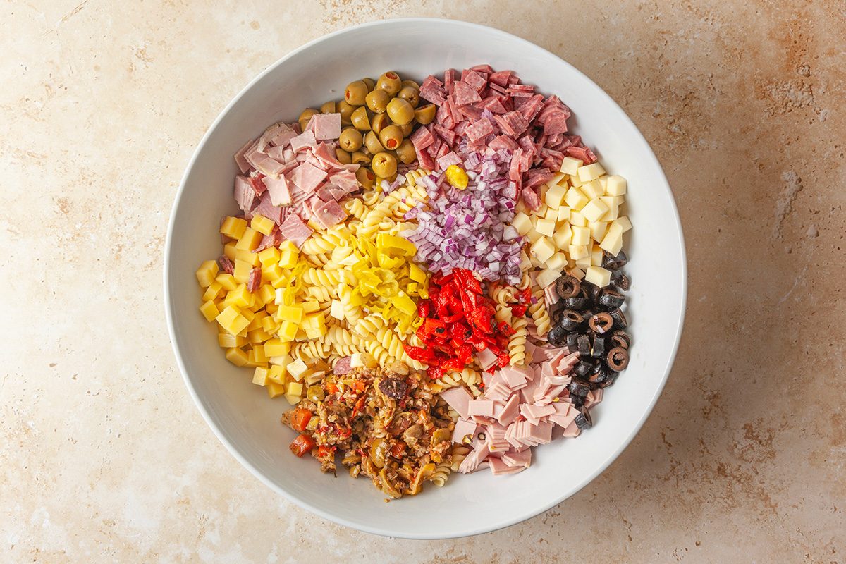 overhead shot of A large white bowl filled with separated piles of cooked pasta, diced meats, cubed cheeses, chopped onions, sliced olives, red peppers, green olives, and a vegetable mixture, all ready to be mixed into a salad