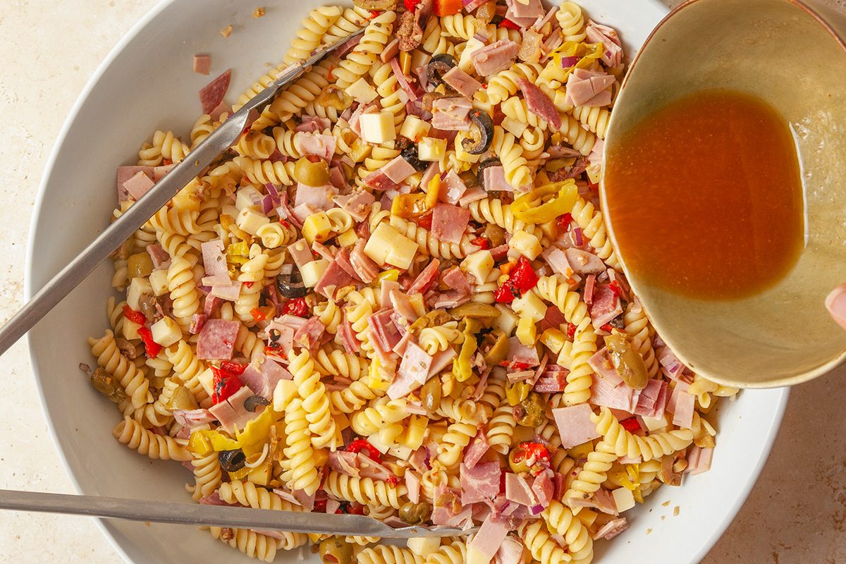 overhead shot of a large bowl of rotini pasta salad with chopped ham, cheese, olives, and peppers, with tongs on the side; A hand is pouring brown dressing from a small bowl onto the salad
