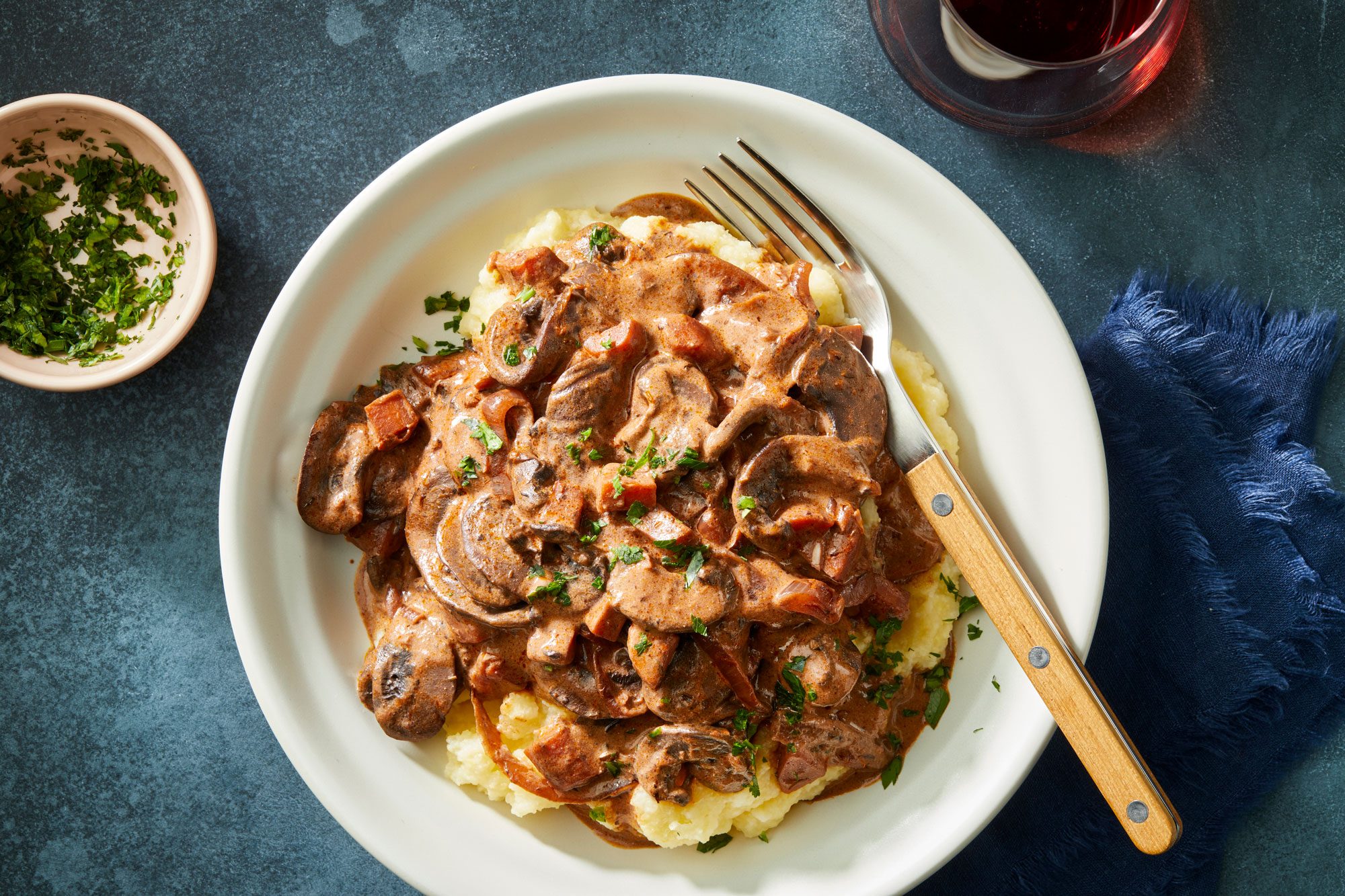 Overhead shot of Mushroom Bourguignon; served in a bowl with a fork and a glass of drink; garnish with parsley; blue napkin; dark blue surface;