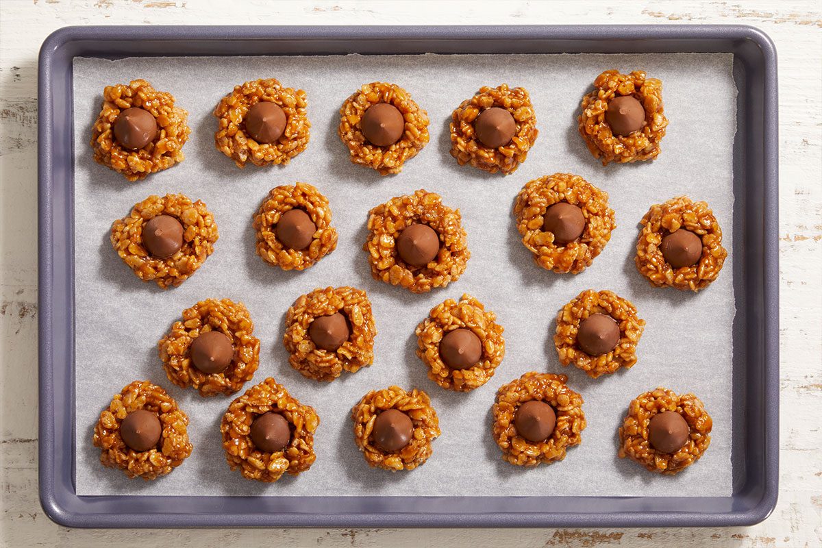 A baking tray lined with parchment paper holds 18 oat cookies, each topped with a round chocolate candy in the center, arranged neatly in rows.