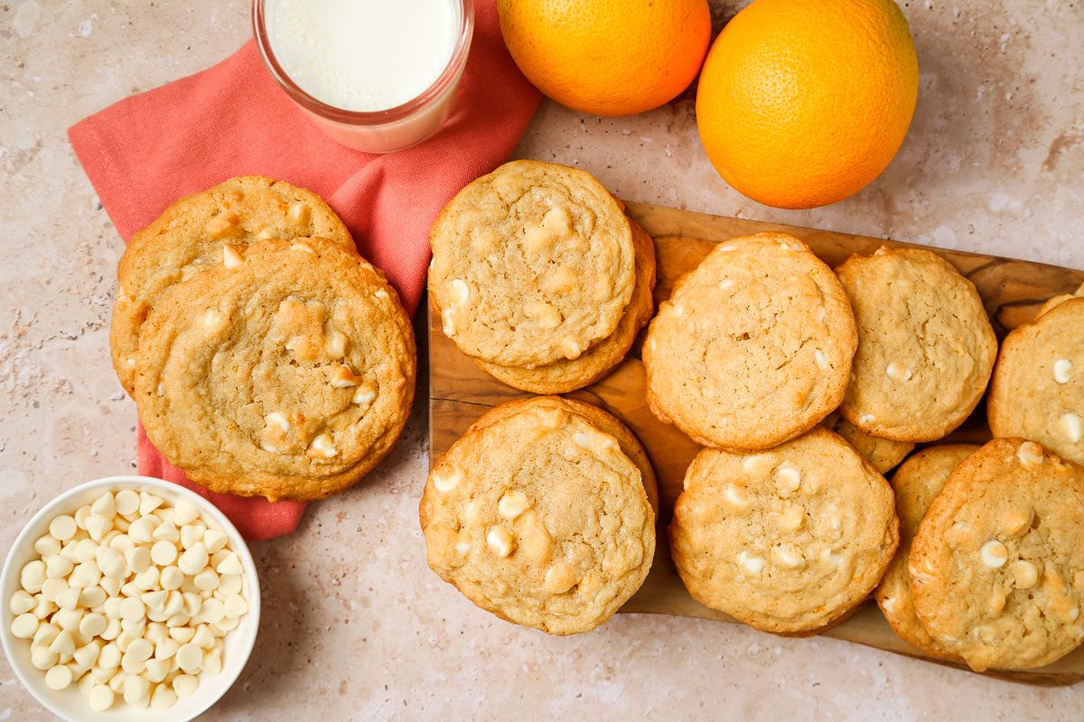 Overhead shot of Orange Creamsicle Cookies served on a wooden tray, with an orange cookie and a glass of milk resting on a napkin; A bowl of white chocolate chips and fresh oranges are placed nearby; all set on a textured marble surface;