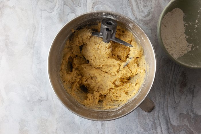 A mixing bowl with cookie dough being mixed by a stand mixer attachment, next to a green bowl containing flour, both on a light gray countertop.