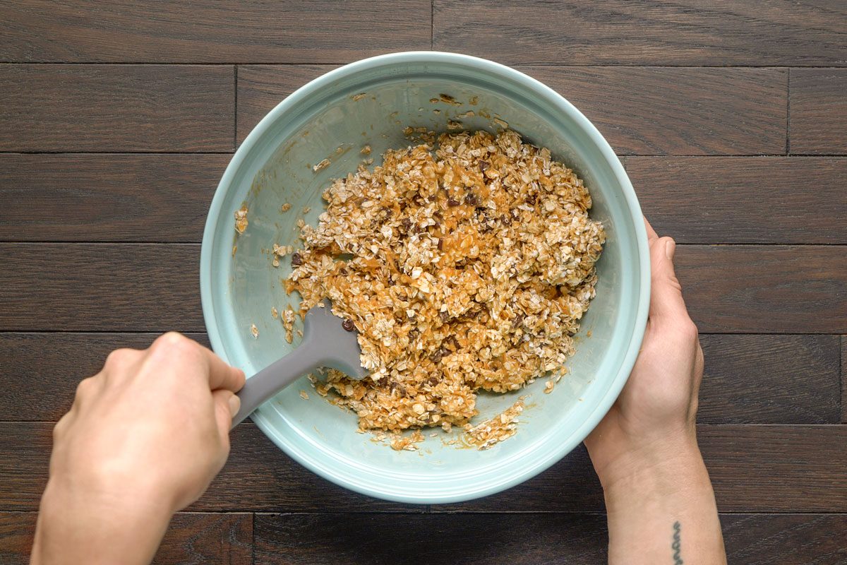 Overhead shot of a person mixing oats with other ingredients in a light blue bowl using a gray spatula; set on a dark wooden surface;