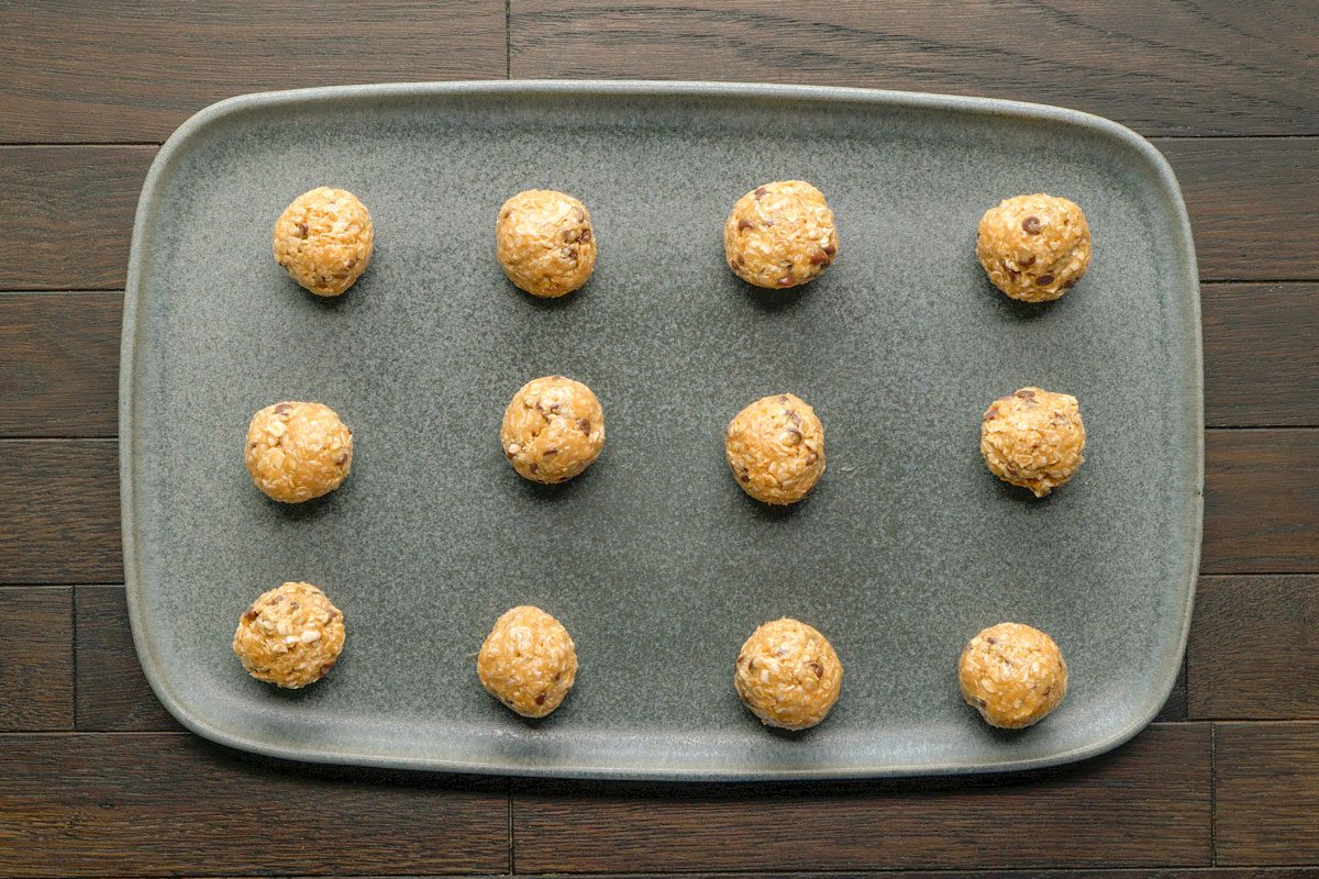Overhead shot of a rectangular gray tray with twelve evenly spaced oatmeal cookie dough balls; arranged in three rows of four; on a dark wooden surface;