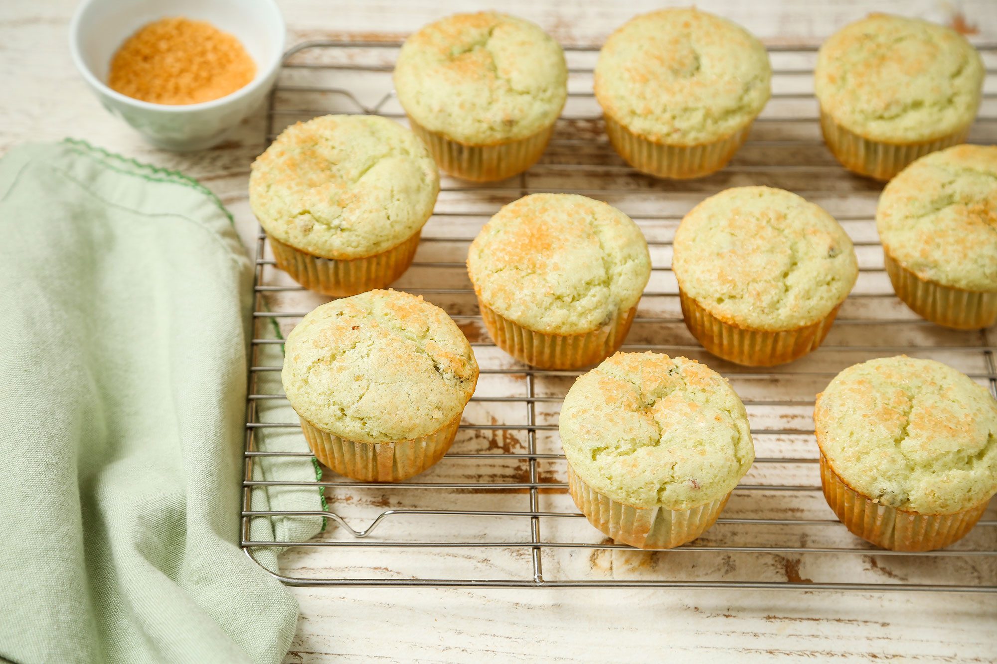 3/4th shot of Ten golden muffins with a light green tint are cooling on a wire rack next to a green cloth, A small white bowl with brown sugar sits in the background on a rustic wooden surface