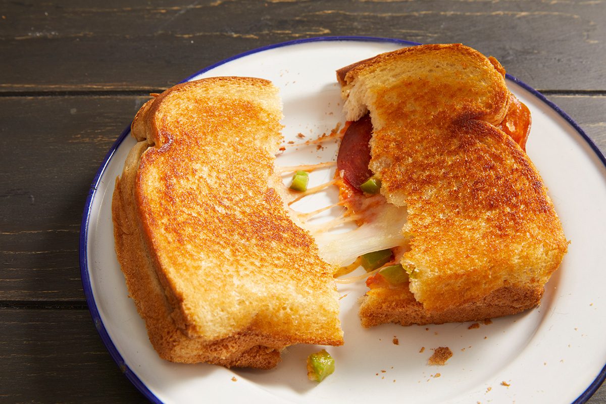 A grilled cheese sandwich with melted cheese, green peppers, and pepperoni is pulled apart on a white plate with a blue rim, revealing the gooey filling inside.