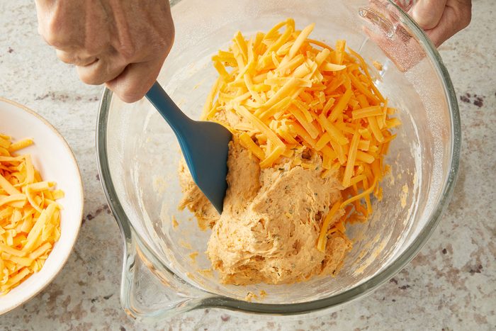 Grated cheese rolled in sticky dough using a rubber spatula in a glass bowl.