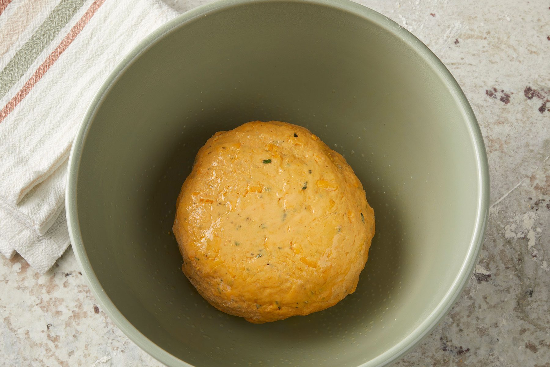 Greased kneaded dough ball in an olive green bowl with a kitchen towel placed on side.