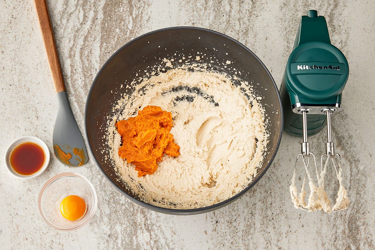 Overhead shot of a mixing bowl with creamed butter and sugar, pumpkin puree on top; nearby are a spatula, a small bowl with an egg; a bowl with vanilla, and a green KitchenAid hand mixer with beaters;