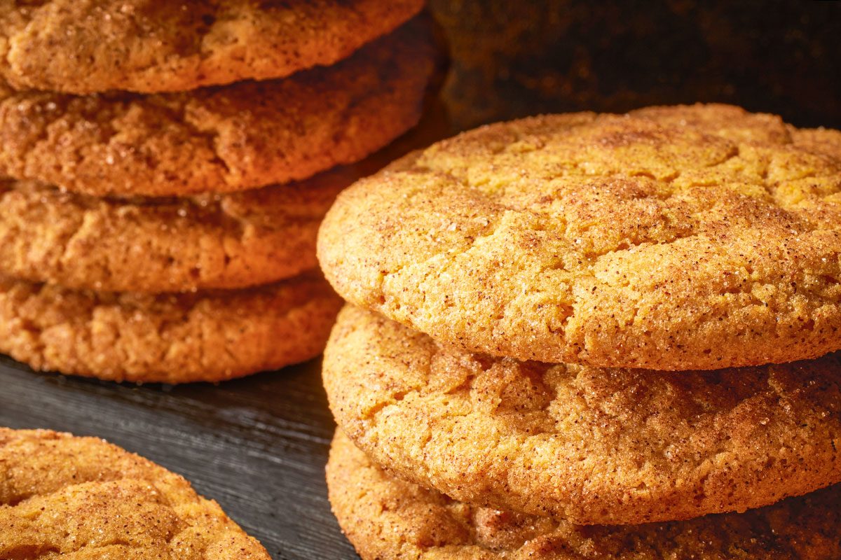 Close-up shot of pumpkin snickerdoodles, stacked and scattered on a dark slate surface; with golden-brown tops coated in cinnamon sugar;