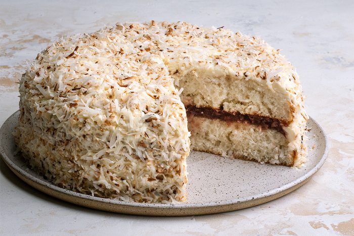 A round coconut cake with shredded coconut covering the frosting, displayed on a ceramic plate. A slice has been cut out, revealing a jam or fruit filling between two layers.