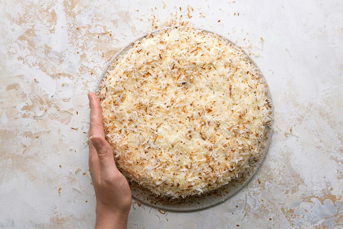 A hand touches the side of a round cake topped with toasted shredded coconut, sitting on a light-colored surface.