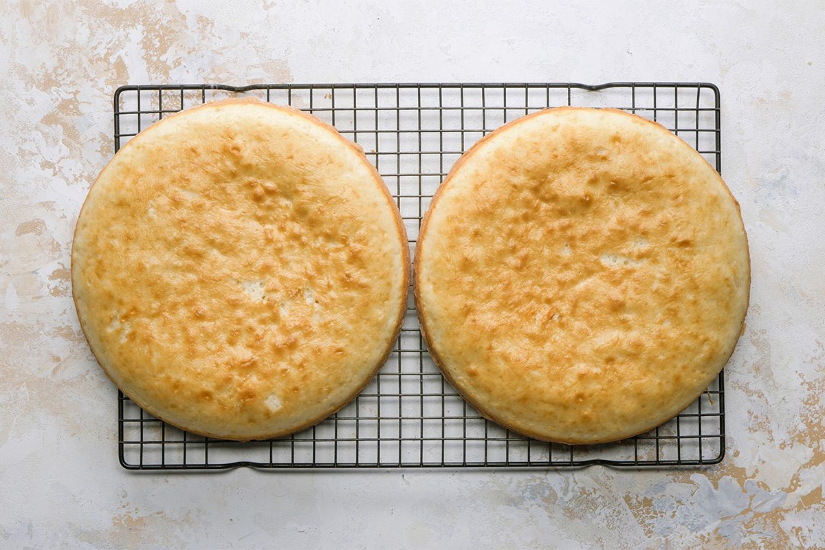 Two round, golden-brown cake layers cooling on a wire rack, placed on a light-colored, textured surface.