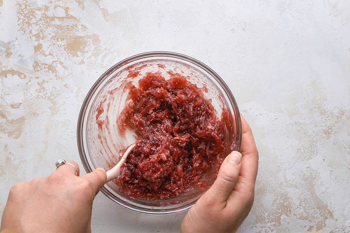 A person stirs a thick, red mixture in a glass bowl with a white spatula on a light-colored surface.