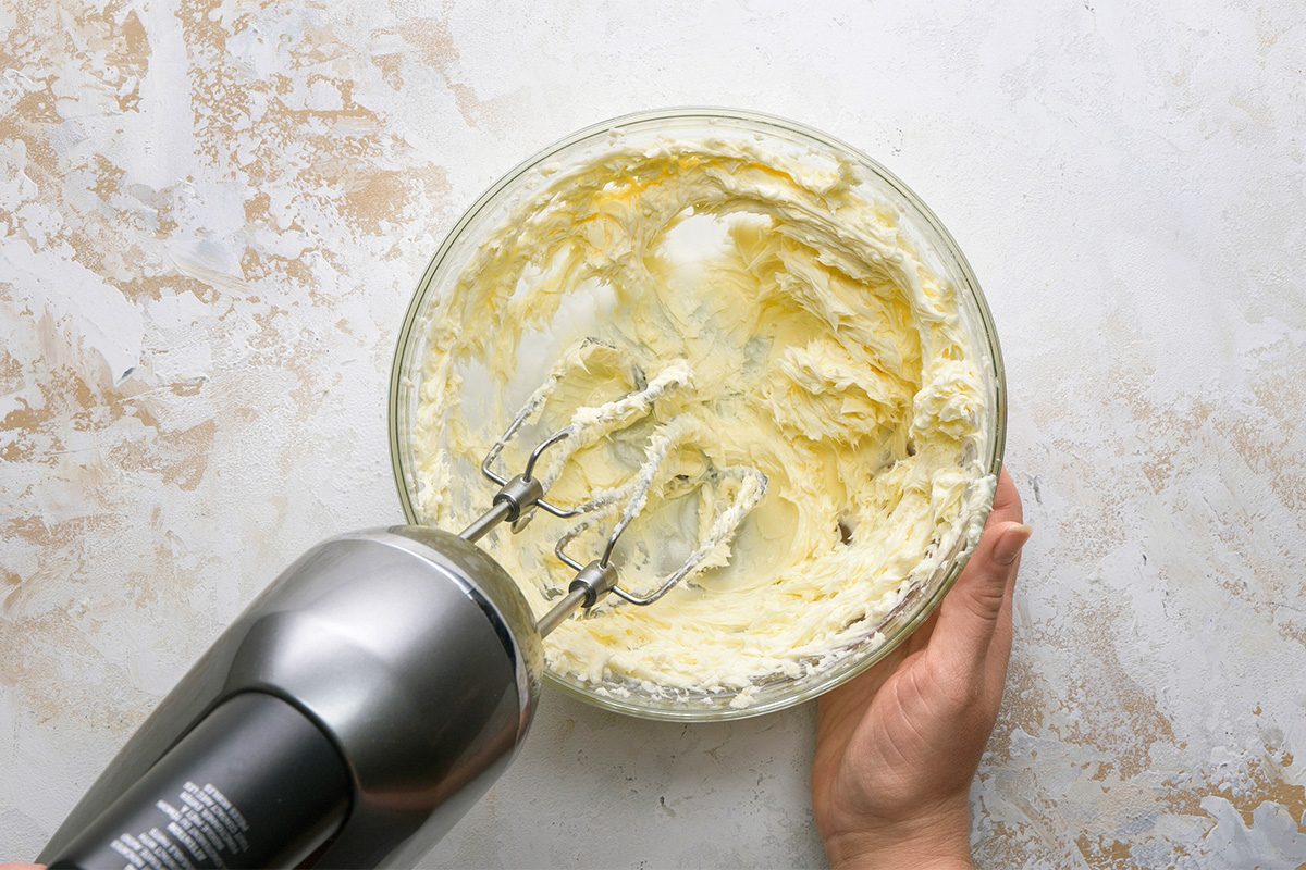 A hand holds a glass bowl filled with creamed butter, while an electric hand mixer with beaters is mixing it against a light, textured surface.