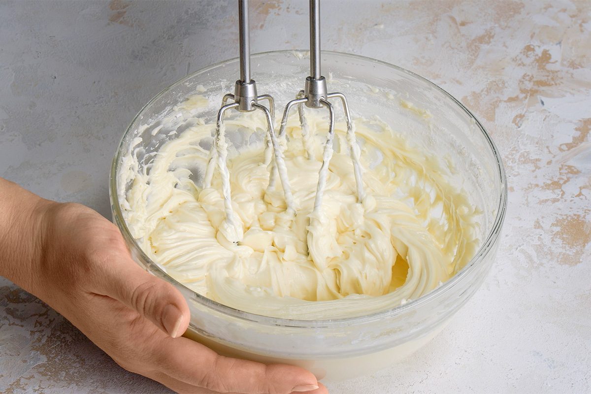 A hand holds a glass bowl filled with creamy batter while an electric hand mixer blends the mixture. The scene is set on a light-colored, textured surface.