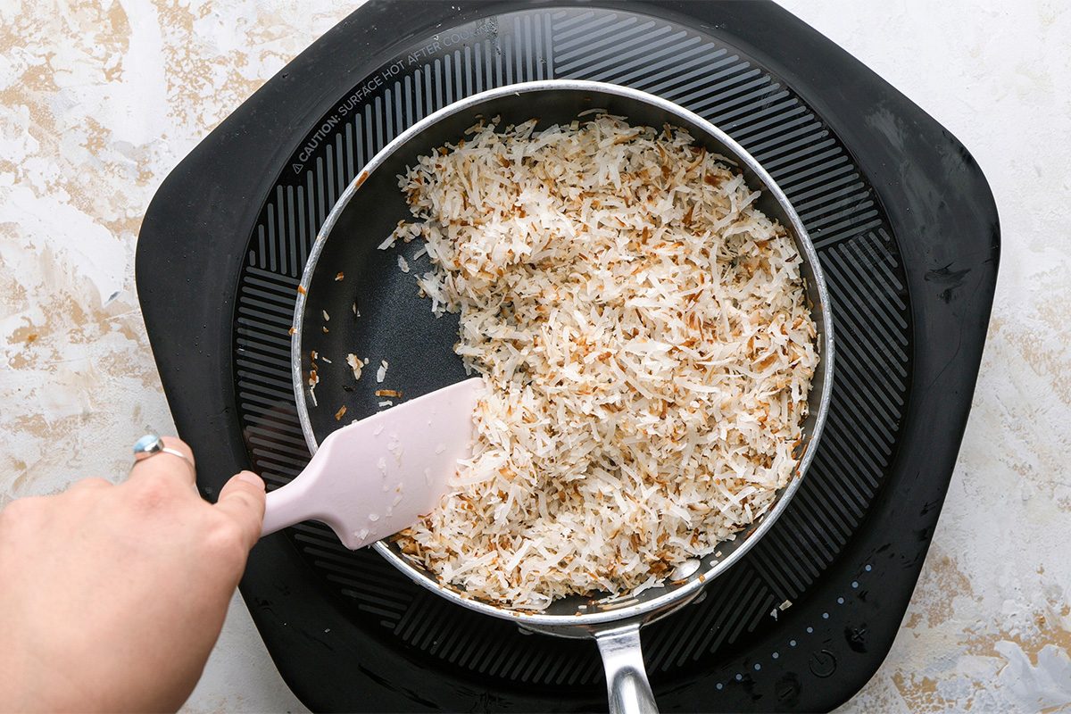 A hand uses a spatula to stir shredded coconut, some of which is toasted, in a pan on a stovetop with a black silicone mat underneath.