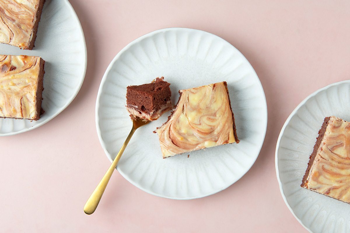 Three white plates, each with a square piece of marbled cheesecake brownie on them, sit on a pale pink surface. One piece has a bite taken out and a fork with a brownie piece is placed on the plate.