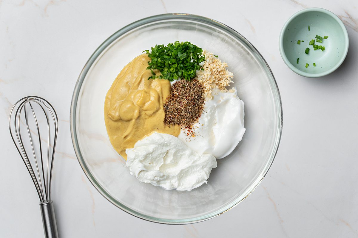 A glass bowl filled with dollops of mustard, sour cream, chopped chives, minced garlic, and seasoning sits on a white countertop next to a whisk and a small bowl with extra chives.