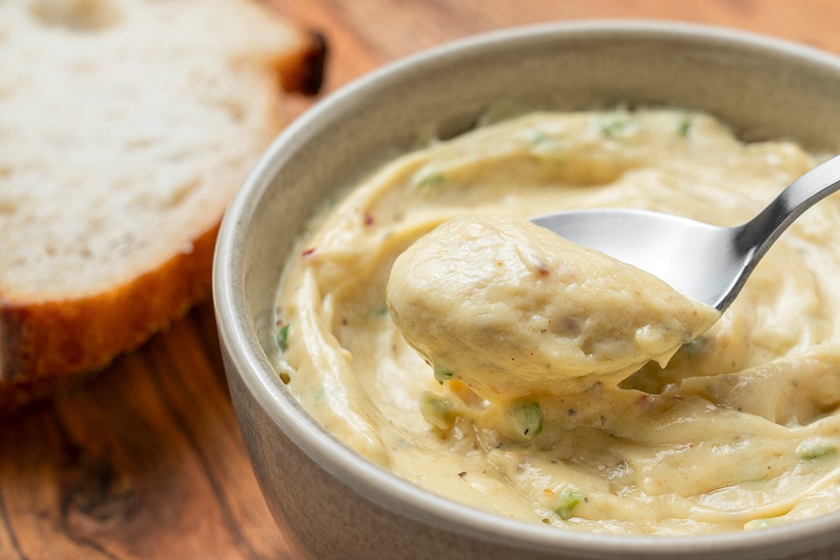 A bowl of creamy, seasoned spread or dip with herbs, being scooped up by a spoon, sits on a wooden surface next to a slice of rustic bread.