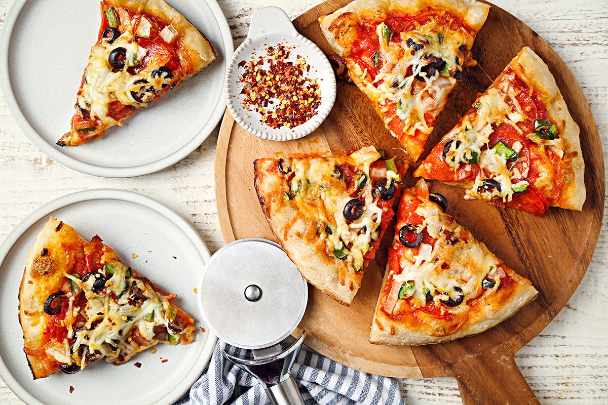 A sliced pizza with cheese, olives, and vegetables sits on a round wooden board. Two slices are served on white plates, with a pizza cutter and a small bowl of red pepper flakes nearby.