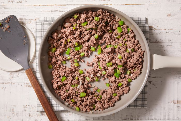 A white skillet filled with cooked ground beef and chopped green bell peppers sits on a plaid cloth atop a white wooden surface. A spatula with a wooden handle rests beside the pan.