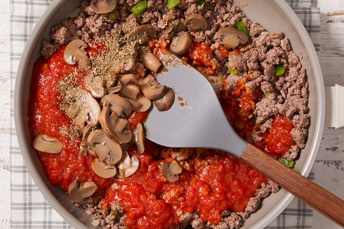 A skillet contains ground beef, tomato sauce, sliced mushrooms, chopped green peppers, and herbs, being mixed with a gray spatula. The skillet rests on a plaid cloth.