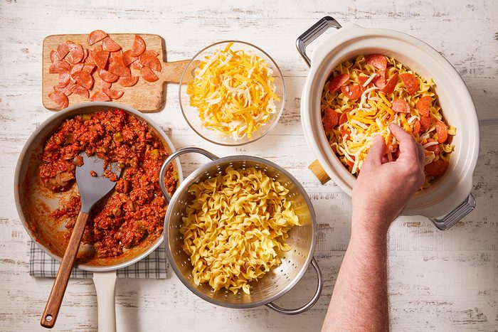 A person assembles a pasta bake, layering cooked pasta, ground meat sauce, shredded cheese, and sliced pepperoni in a baking dish, with ingredients and utensils arranged on a white wooden surface.