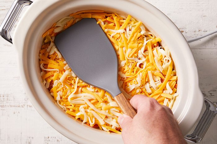 A hand holding a gray spatula over shredded cheese in a white slow cooker on a light-colored surface.