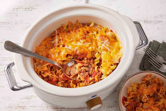 A slow cooker filled with cheesy noodle casserole, with a serving spoon inside and a portion scooped into a small bowl beside it. The casserole is topped with melted cheese.