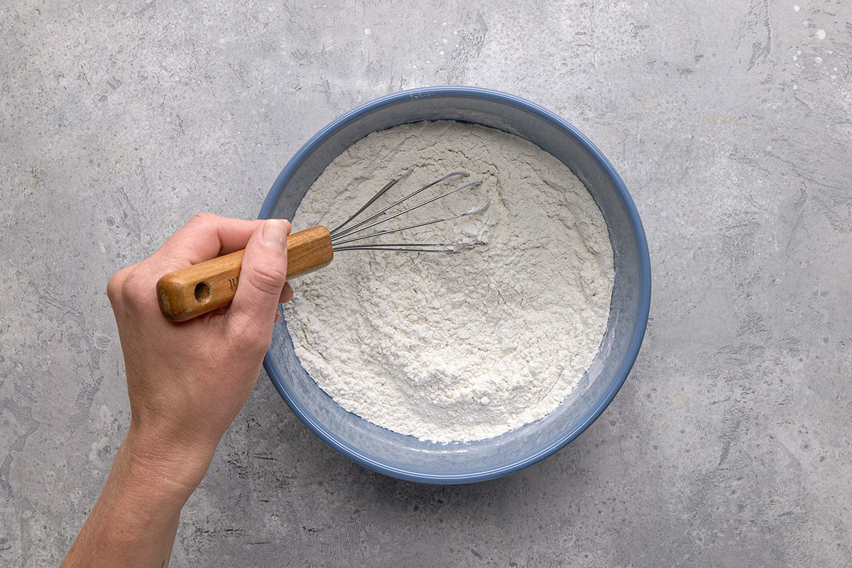 A hand holding a whisk stirs flour in a blue bowl on a gray surface.