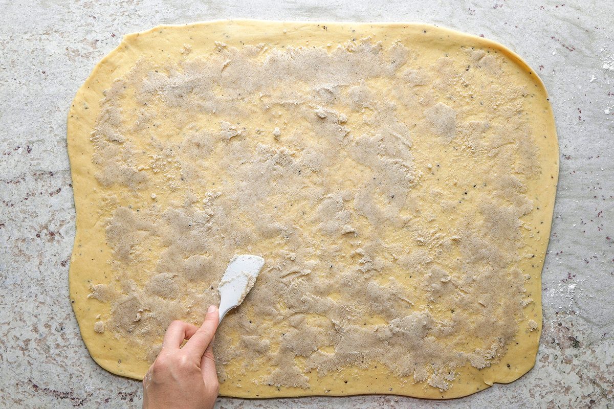 overhead shot of A hand spreads a layer of butter and cinnamon sugar onto a large rectangle of rolled dough on a light, floured surface