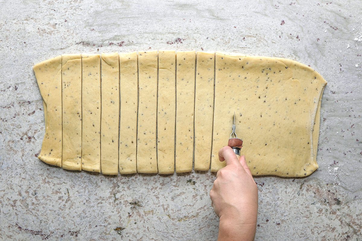 overhead shot of A hand uses a pastry cutter to slice a rectangular sheet of speckled dough into even strips on a lightly floured surface