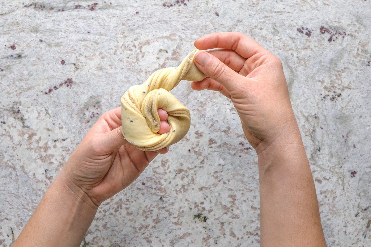 overhead shot of Two hands twist a strip of dough into a knot shape on a light, speckled countertop
