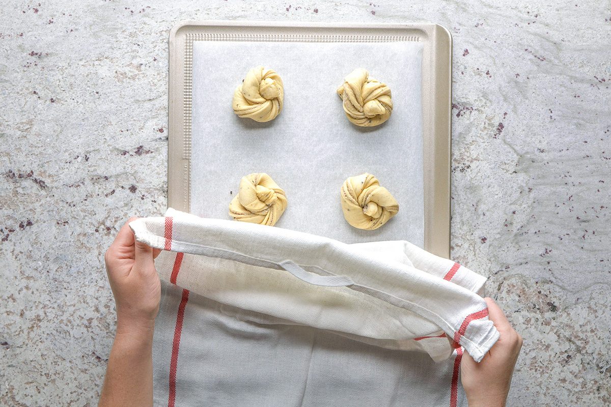 overhead shot of Hands covering a baking sheet with four unbaked twisted dough buns using a white cloth with red stripes, on a light countertop