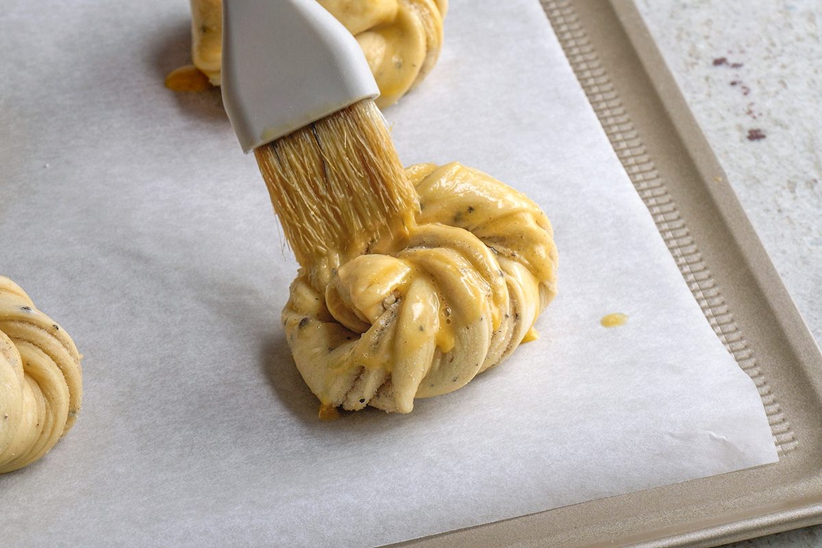 closeup shot of a pastry brush applying egg wash to a twisted knot of dough with visible herbs, placed on a parchment lined baking sheet