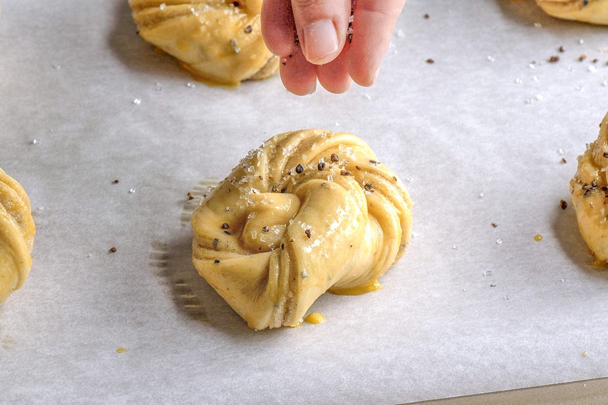 closeup shot of A hand sprinkles coarse salt and seeds onto a twisted, unbaked pastry dough placed on parchment paper, ready for baking; Other similar dough pieces are visible in the background