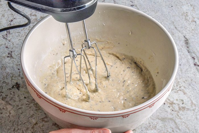3/4th shot of A hand holds a white mixing bowl with red trim, containing thick batter being mixed by an electric hand mixer with two beaters, on a light gray textured surface